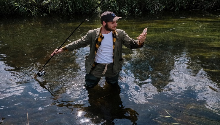 Male standing in lake thigh high looking at bass fish caught on hook holding fishing pole